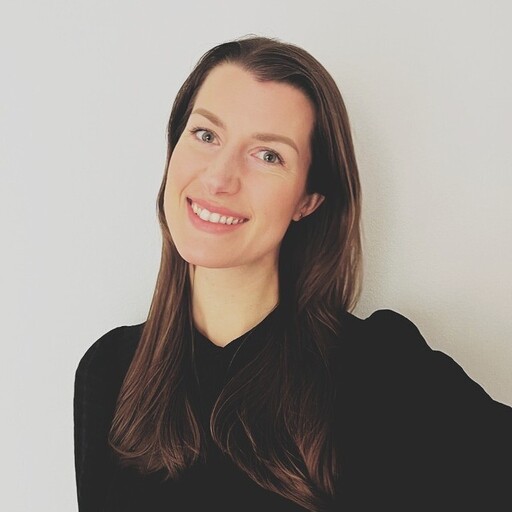 En person med långt brunt hår, klädd i en svart tröja, står mot en ljus bakgrund.