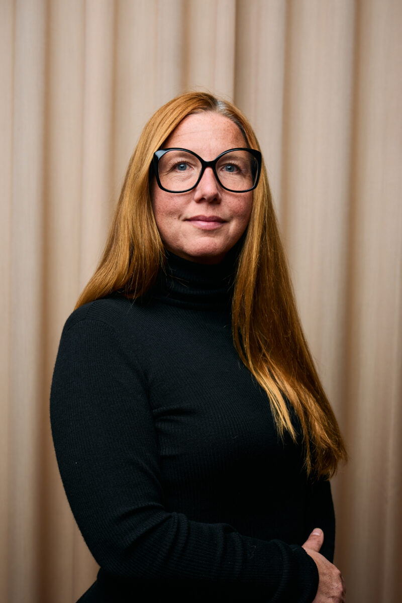 En person klädd i en svart polotröja med långt rött hår, stående framför en ljus gardinbakgrund.