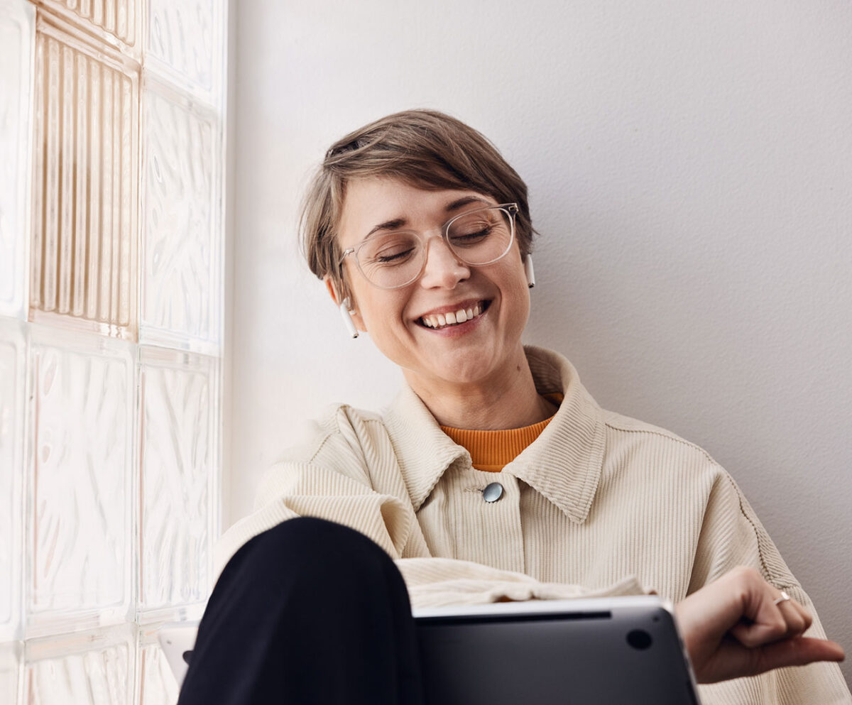 En person i ljus jacka och orange tröja sitter vid ett fönster med frostat glas, håller en surfplatta och har en vit hörlur i örat.