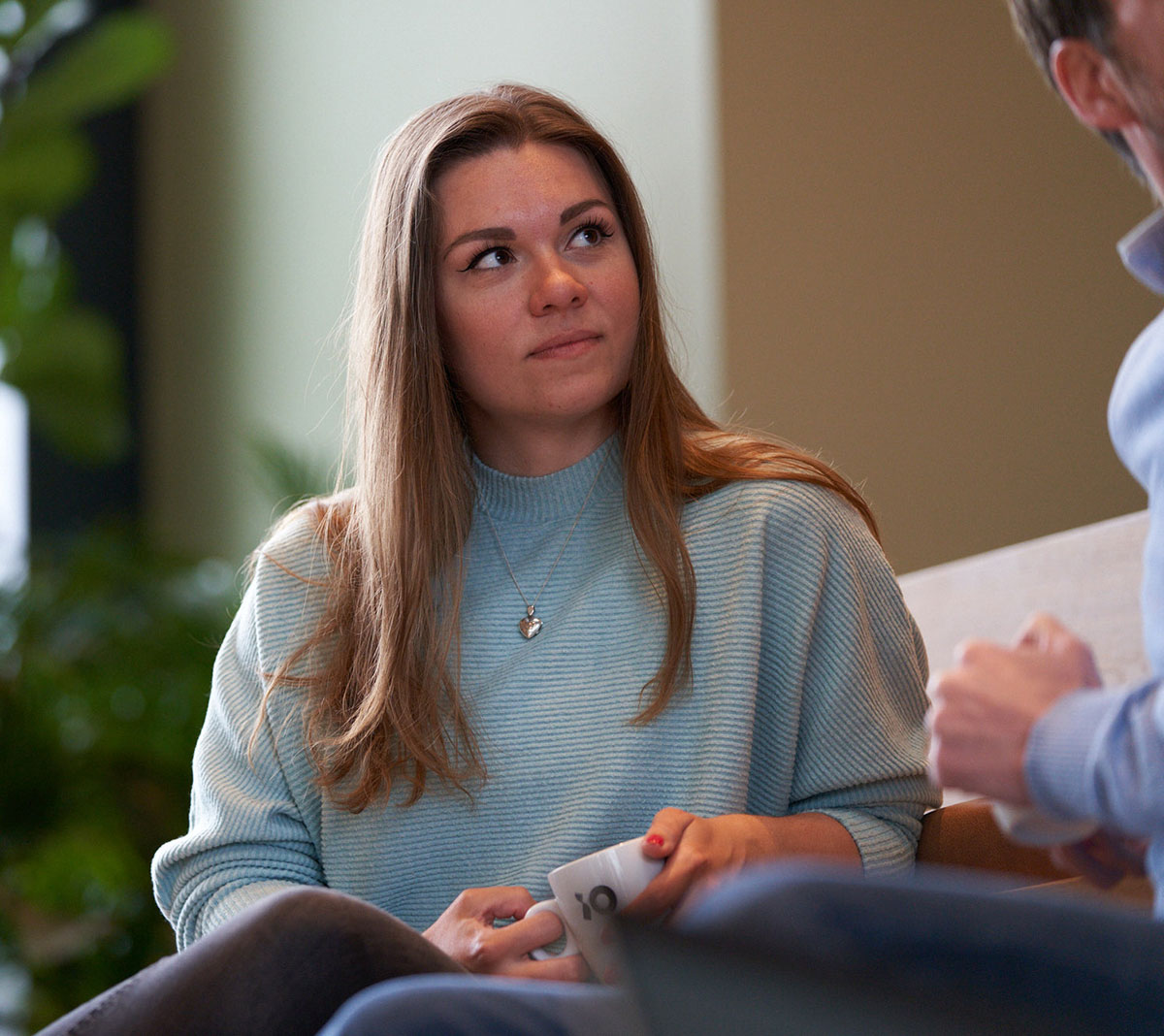 En person med långt hår i en ljusblå tröja håller en vit kopp med texten "jo" i en avslappnad miljö med grönska i bakgrunden.