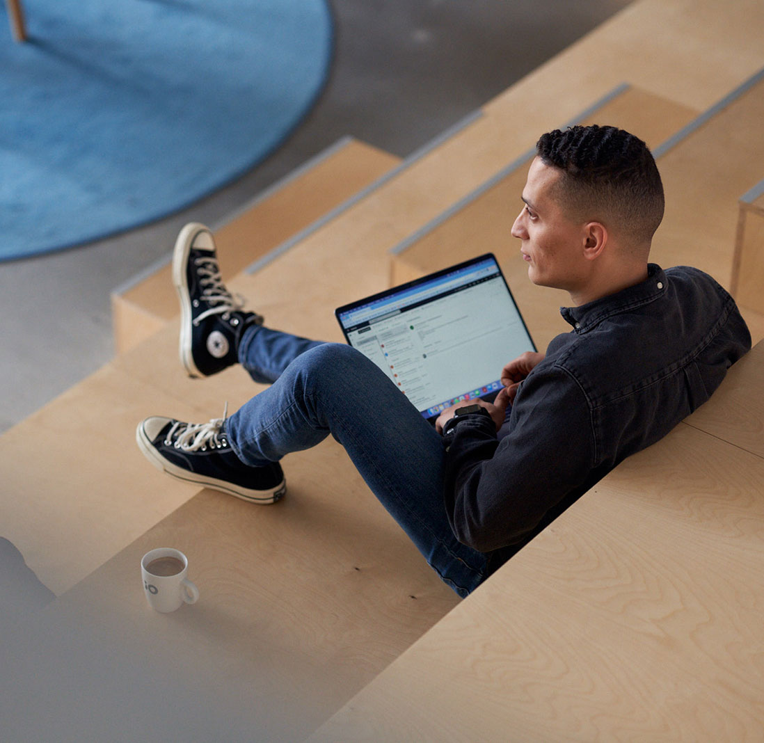 En person sitter på en trätrappa med en bärbar dator i knät, en kaffekopp står på trappan bredvid.