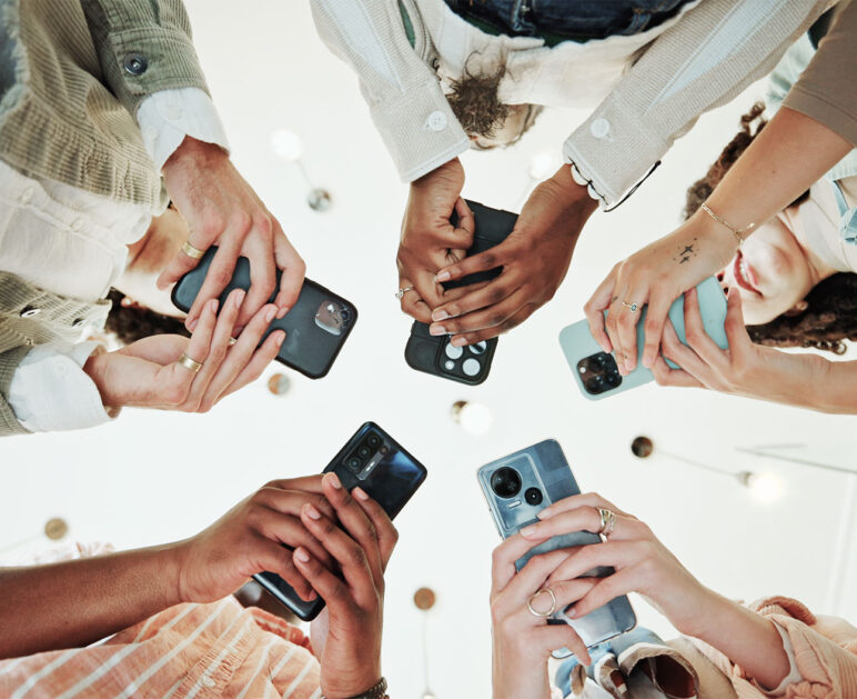 En grupp människor håller upp sina mobiltelefoner mot kameran, med varierande telefonmodeller och färger, sett från ett nedifrån-perspektiv.