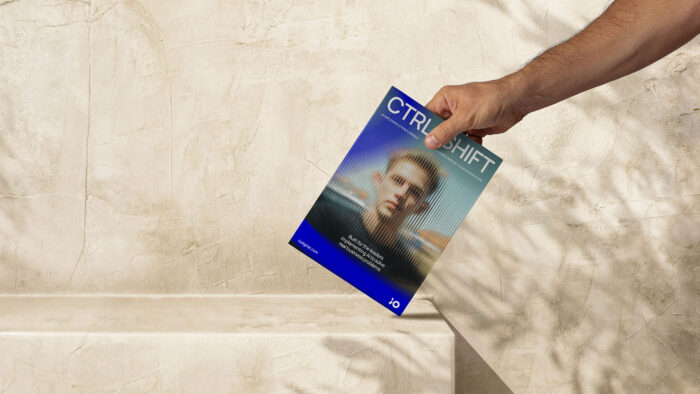 En hand håller en broschyr med titeln "CTRL SHIFT" i blått och vitt mot en beige bakgrund med skuggor. Texten på broschyren lyder: "Built for the leaders implementing AI to solve real business problems" och "bousgual.com".