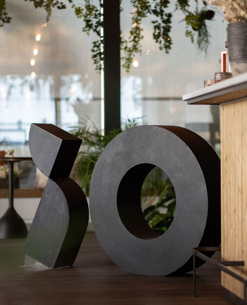 En stor svart skulptur i form av iO står på ett trägolv, omgiven av gröna växter och inredning i en modern miljö.