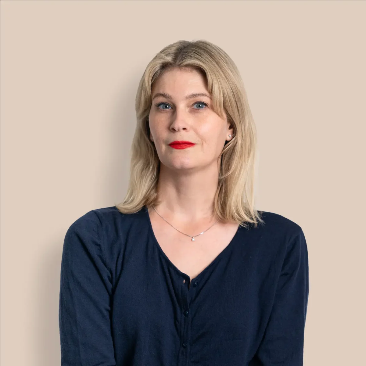 En kvinna med blont hår och rött läppstift, klädd i en mörkblå blus, står framför en beige bakgrund.